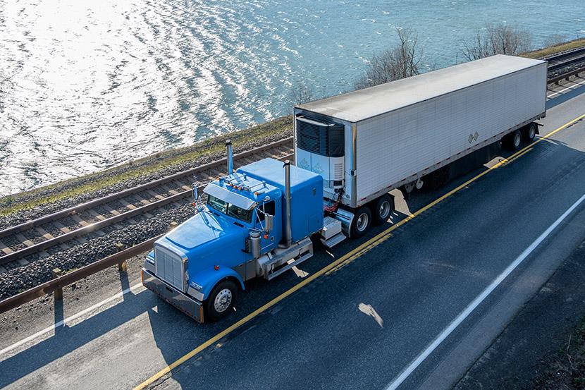 blue big rig semi truck pulling cargo in refrigerated trailer driving on road next to railroad track